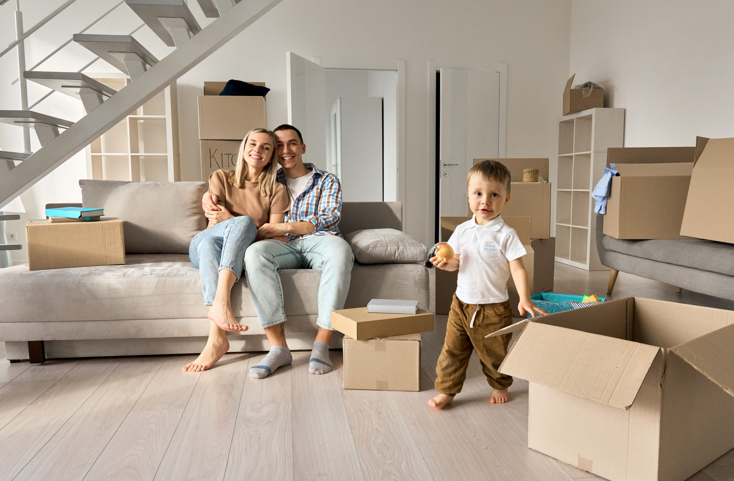 Happy family new home owners with child son in living room on moving day. Young couple apartment buyers relaxing on couch with kid boy playing around boxes. Relocation and house renovation, removal.