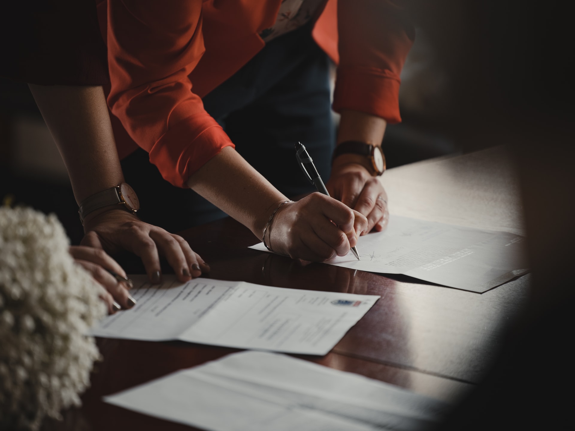closeup of two people's arms and hands cosigning mortgage documents on a wooden table